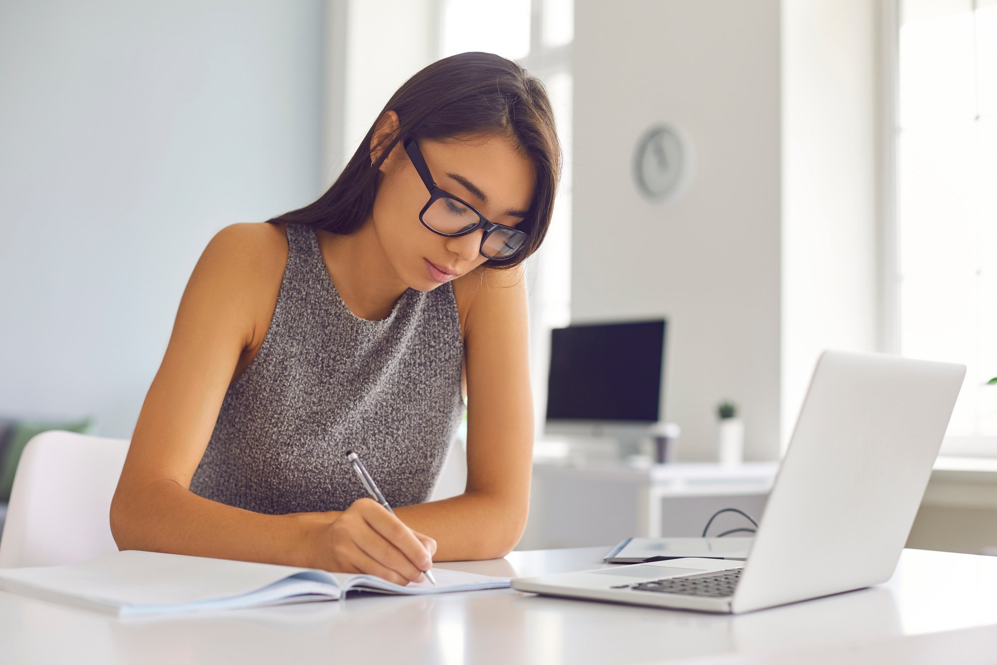 Young Girl Office Worker Sitting at a Desk in Front of a Laptop and Making Notes in a Notebook.